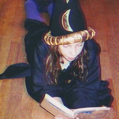 photo of Annie James as a 9- or 10-year old. she is a white girl with long-ish brown hair. she is not looking at the camera; instead, she is reading a book. she is lying on her stomach on the floor and wearing a wizard robe, purple leggings, and a Halloween wizard hat with gold stars and moons.