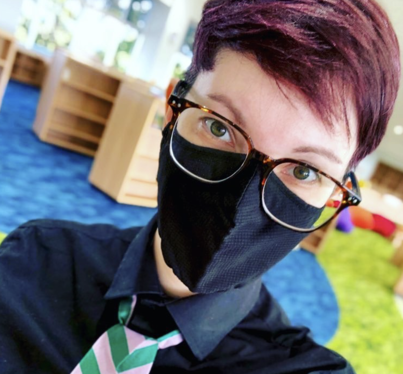 Annie James, a white person with short pink hair, tortoiseshell glasses, and a black N-95 mask, in their library workspace